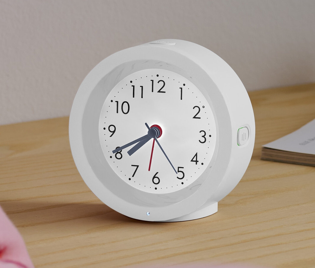Bílý analogový quartzový budík s podsvícením stojí na nočním stolku vedle postele.