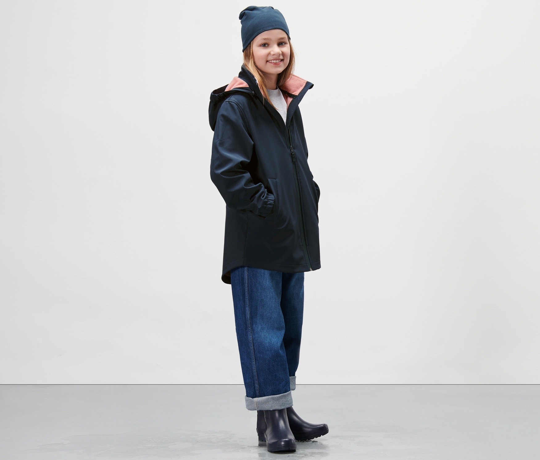 Dívka má na sobě tmavě modrý softshellový kabát, džíny a čepici.