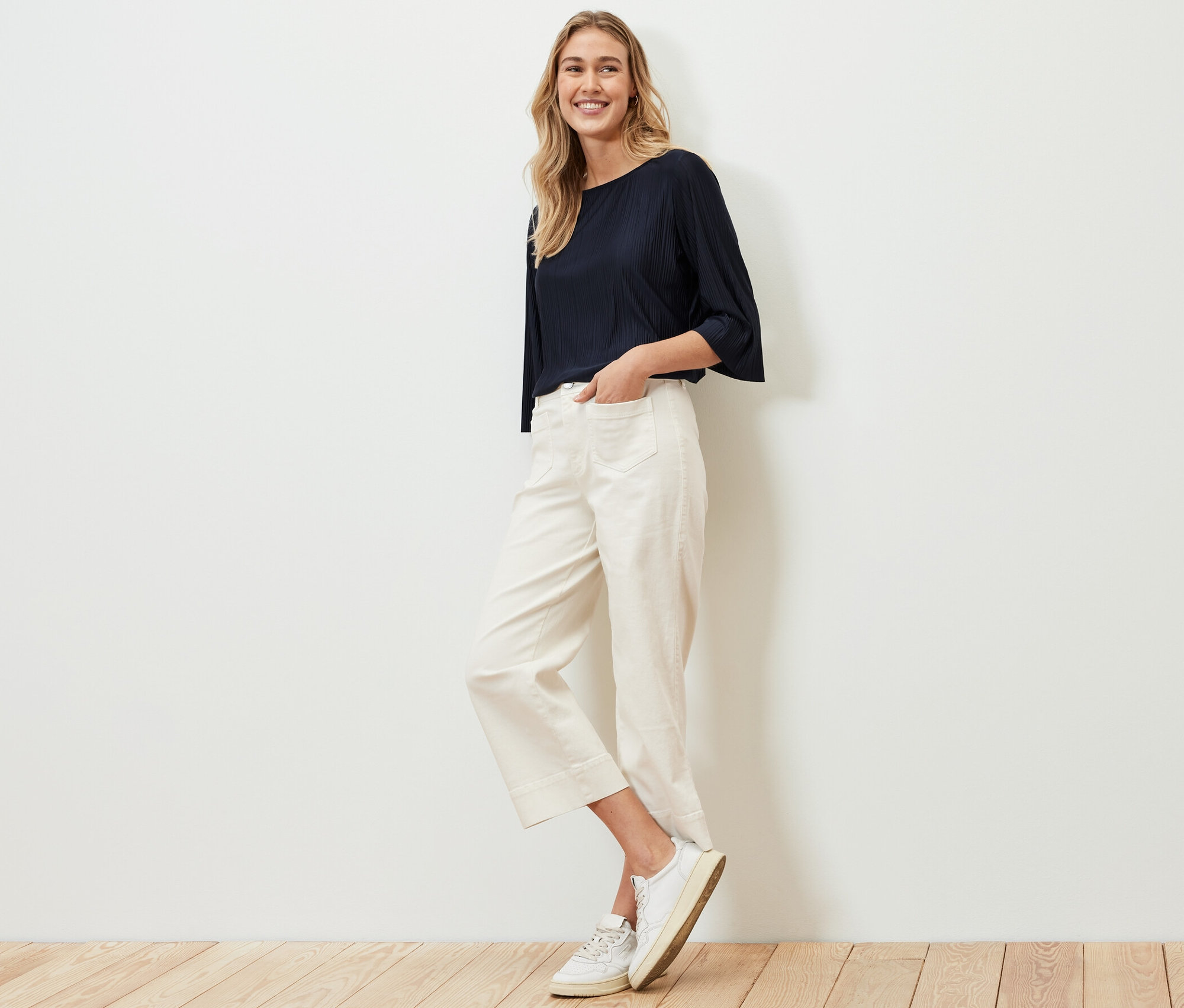 Žena pózuje v plisovaném halenkovém triku, bílých kalhotách a teniskách.