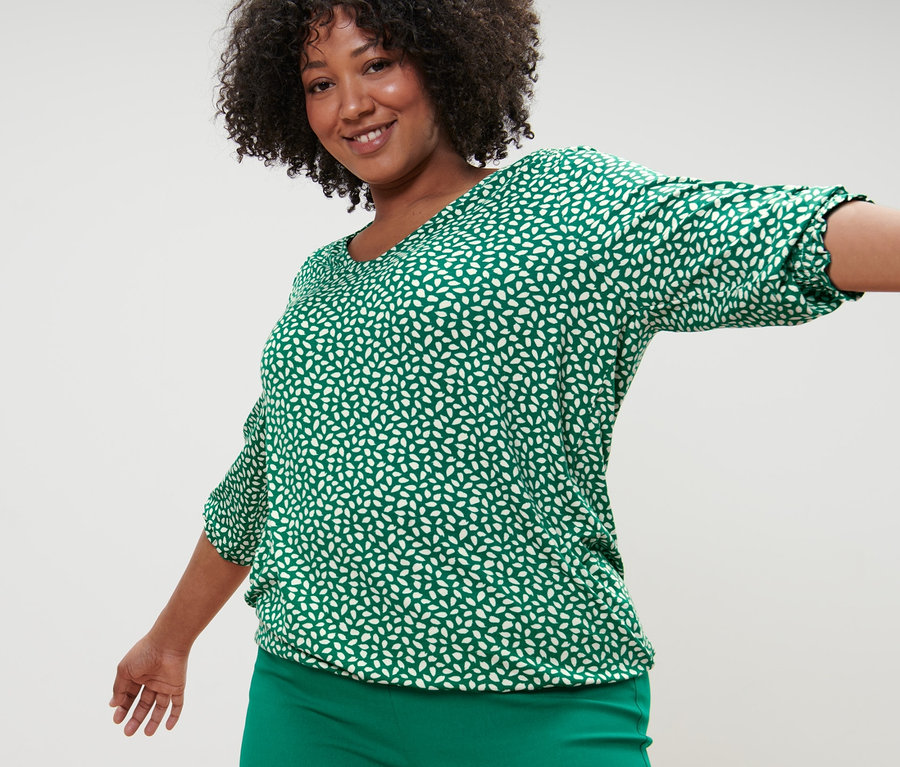 Žena pózuje v zeleném halénkovém triku s potiskem a zelených strečových kalhotách.