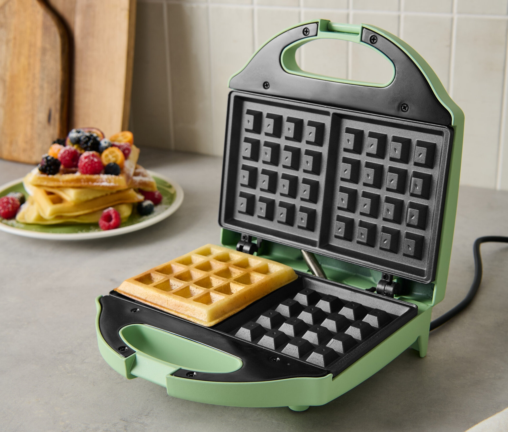 Zelený vaflovač s upečenou vaflí uvnitř. Na talíři leží hromada vaflí s ovocem.