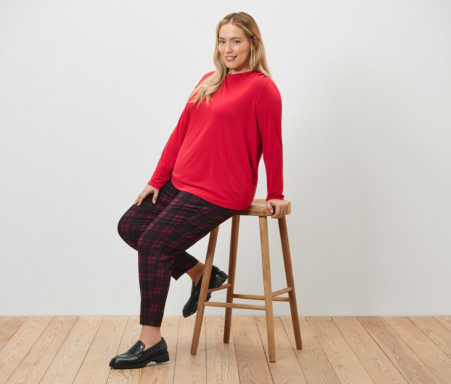 Žena sedí na stoličce a má na sobě kostkované strečové kalhoty a halenkové triko s řasením.