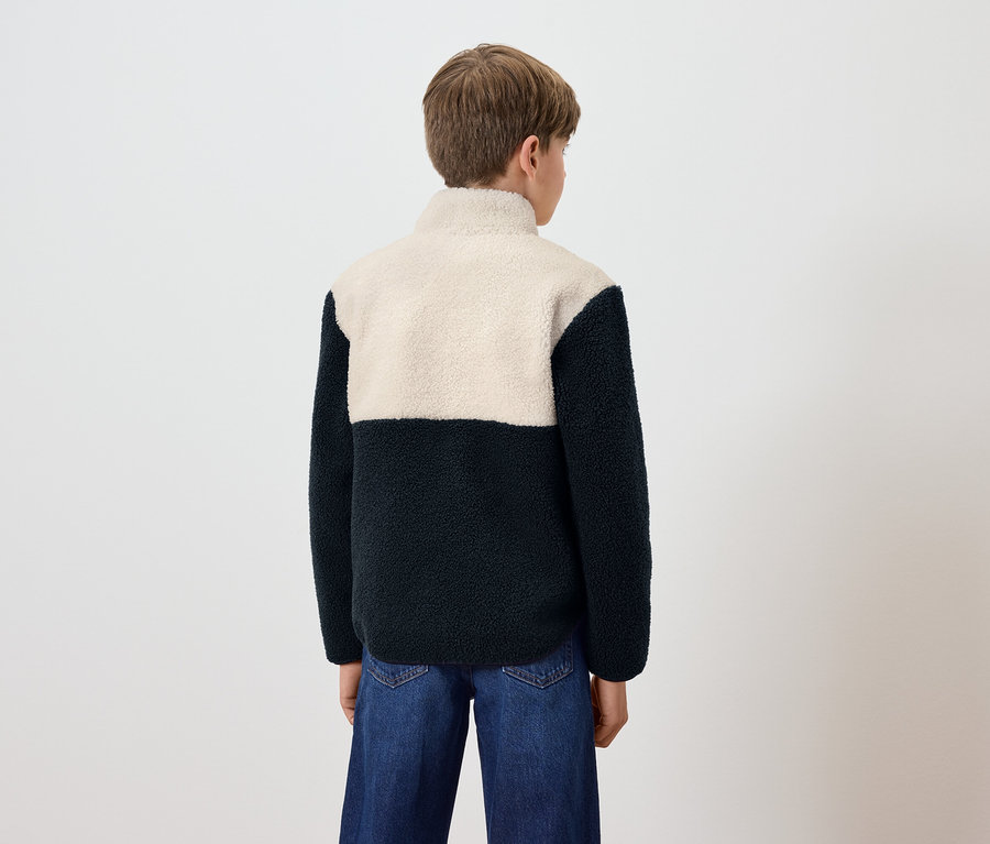 Chlapec zády k fotoaparátu má na sobě modré džíny a fleecovou mikinu s krémovým topem a tmavě modrým spodkem.