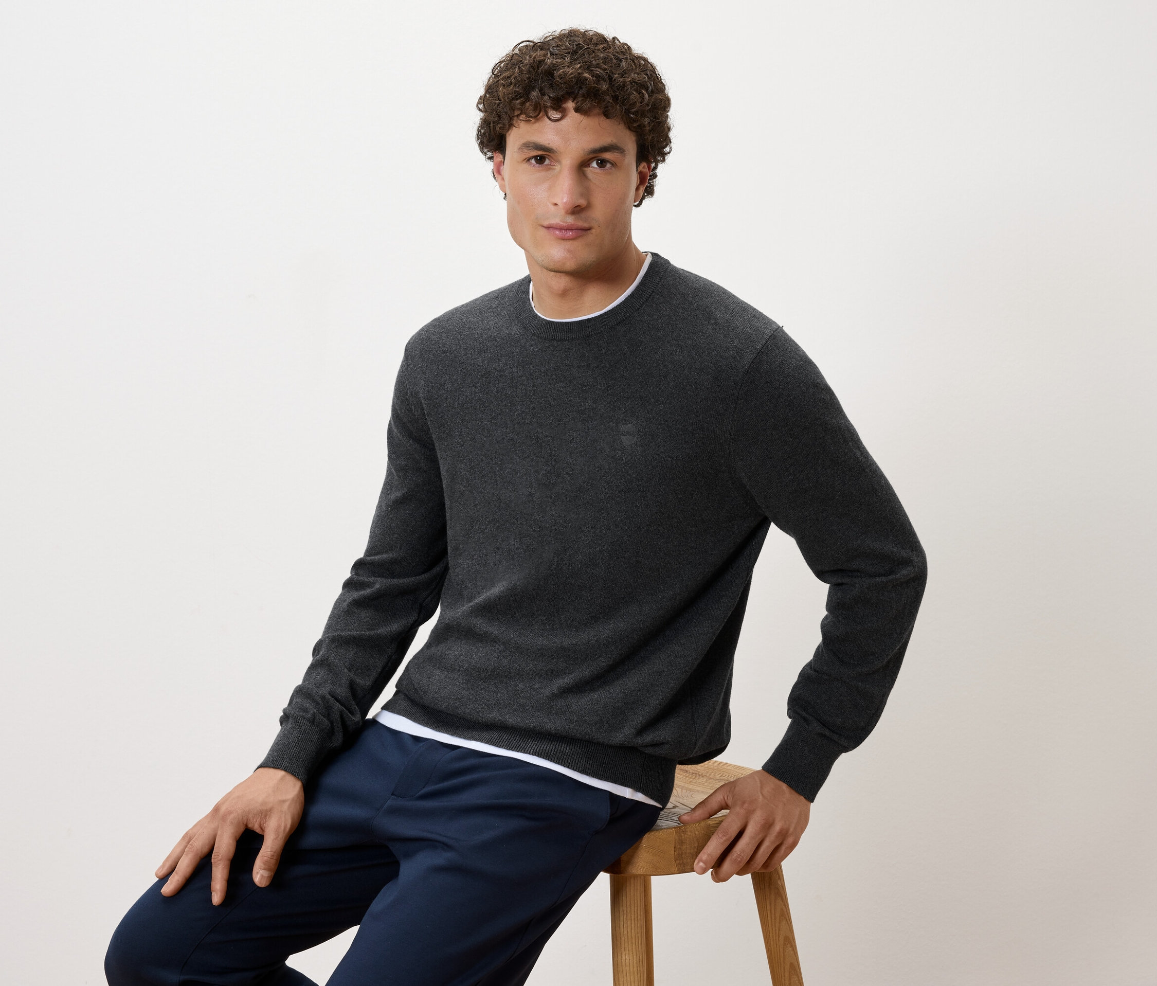 Muž sedí na stoličce, oblečený v šedém svetru a tmavě modrých kalhotách.
