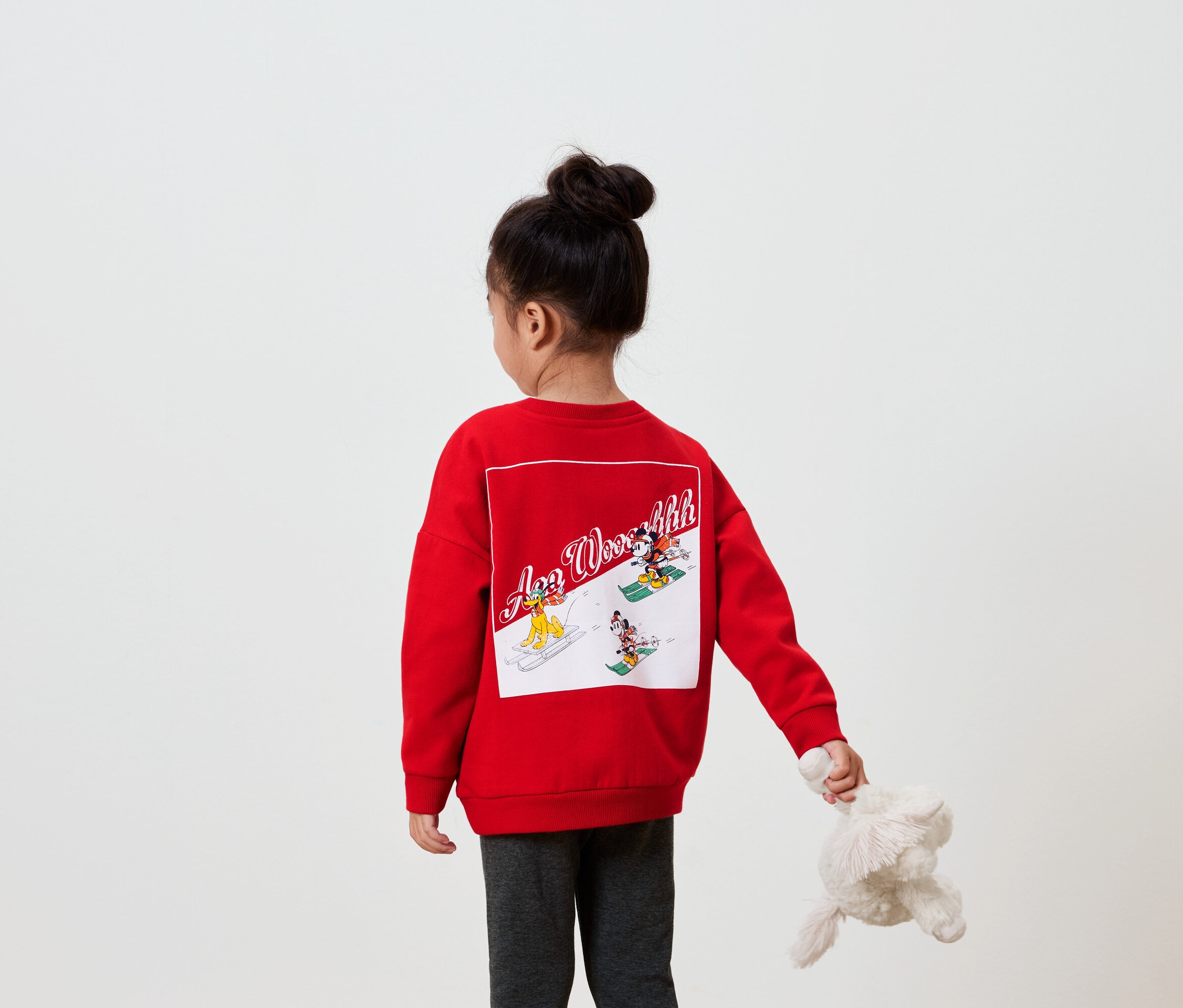 Dívka zády k fotoaparátu má na sobě červenou mikinu s obrázkem na zádech a v ruce drží plyšovou hračku.