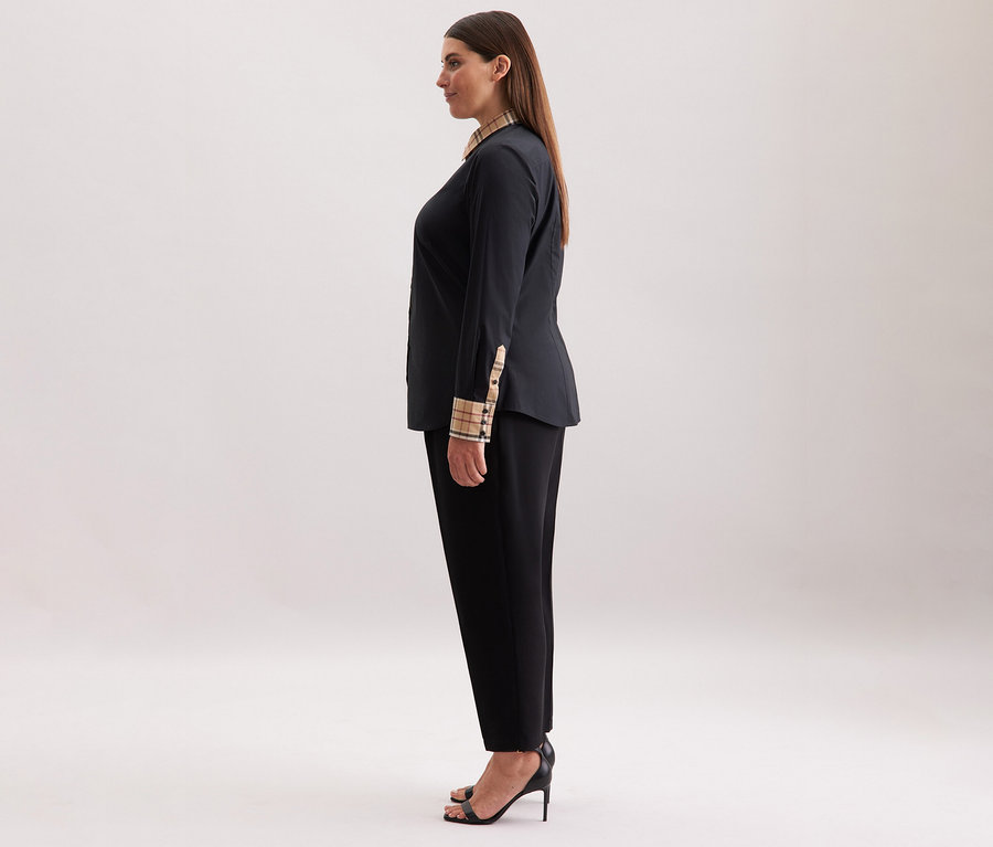 Žena má na sobě černou popelínovou halenku s kostkovaným detailem Seidensticker, černé kalhoty a černé podpatky.