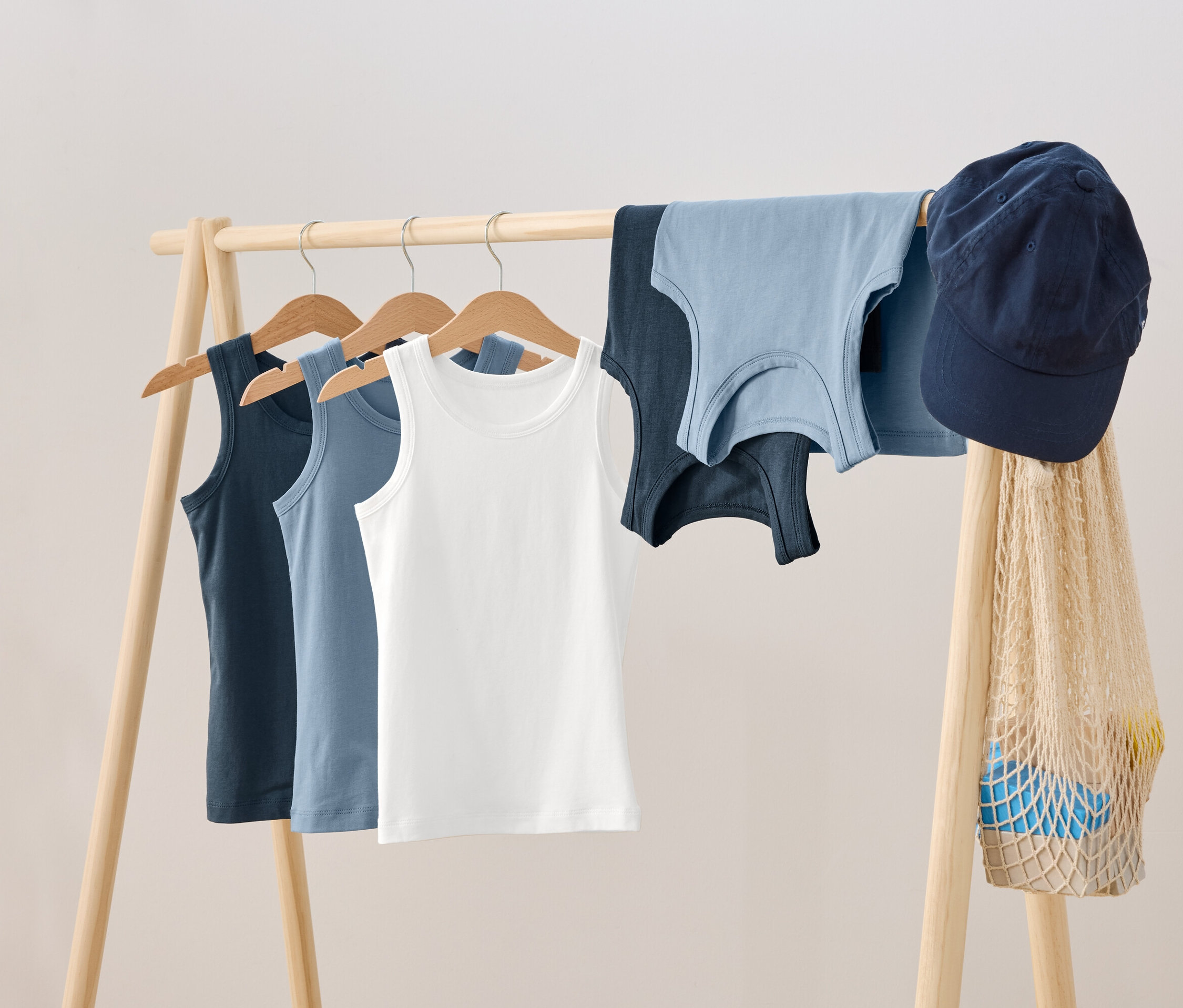 Na dřevěném věšáku visí tři tílka na ramínkách, vedle nich leží dvě další tílka, modrá kšiltovka a síťovaná taška.