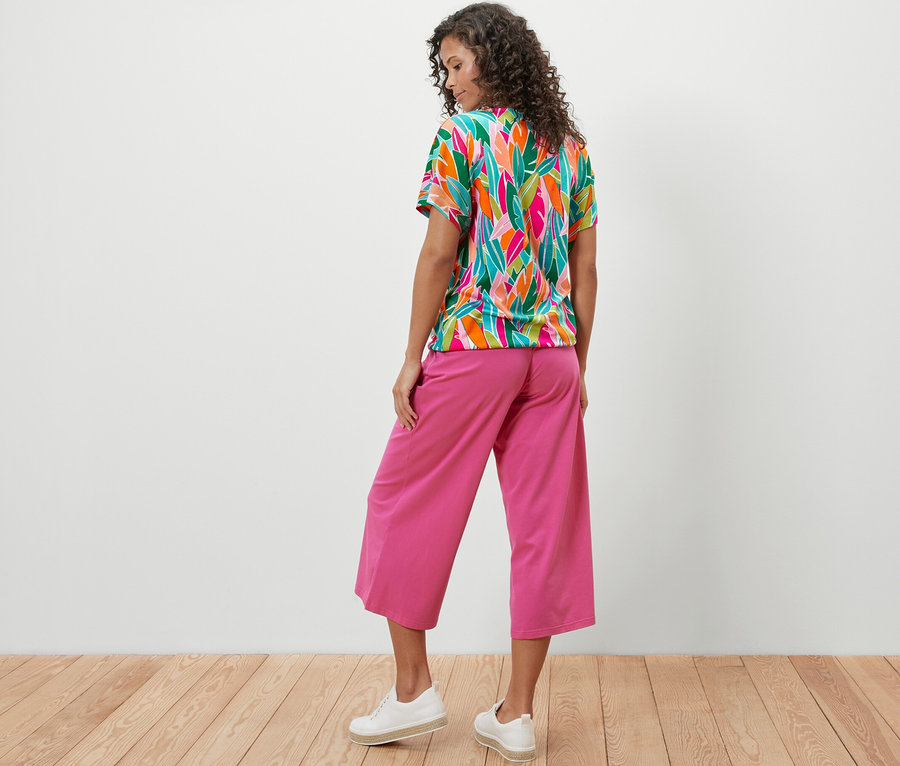 Žena stojí zády k fotoaparátu a má na sobě halenkové tričko se spadlými rameny a celoplošným potiskem a růžové kalhoty.