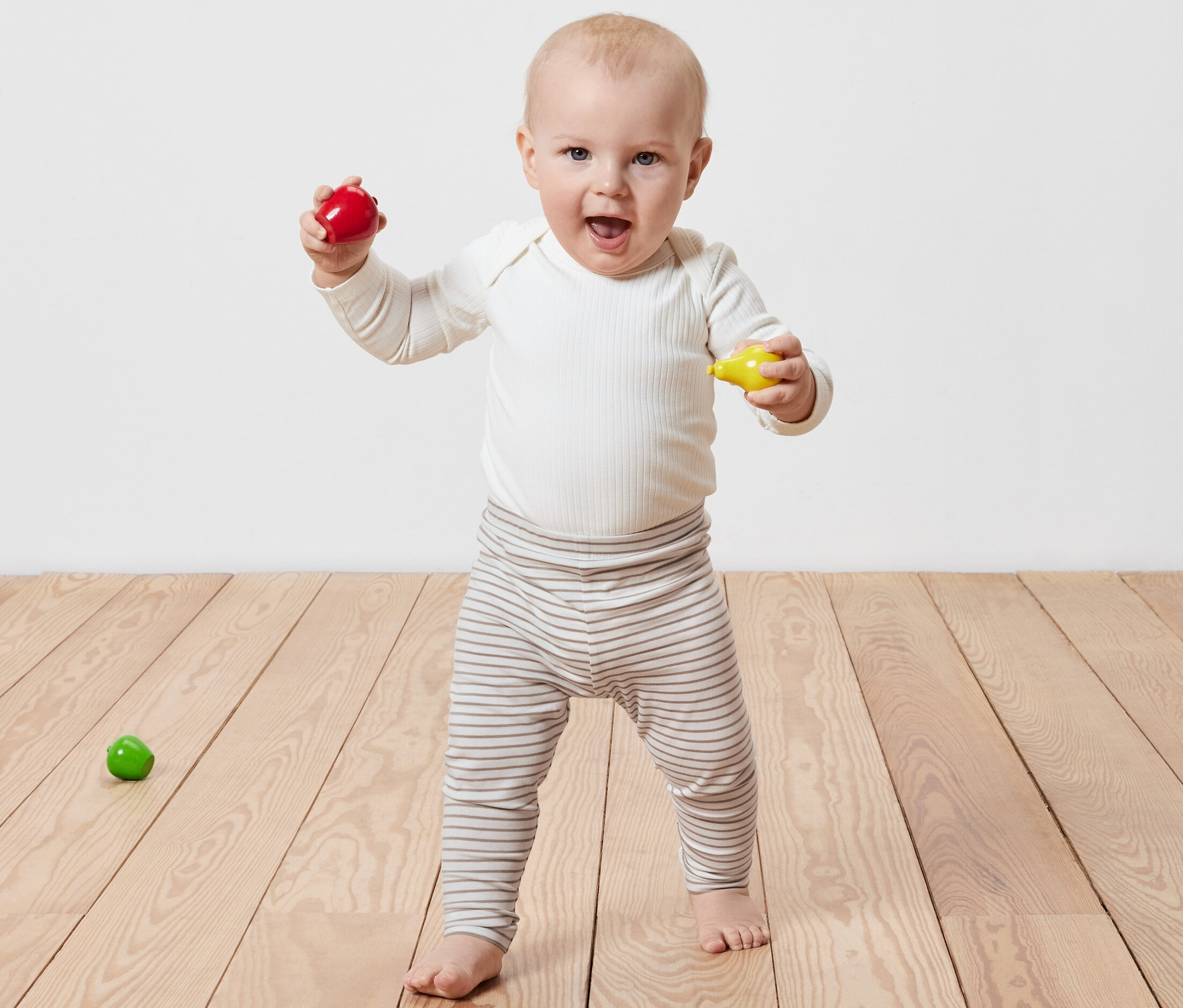 Usmívající se dítě stojí na dřevěné podlaze a drží v rukou červenou a žlutou hračku. Na sobě má legíny Dětské legíny, 5 ks.