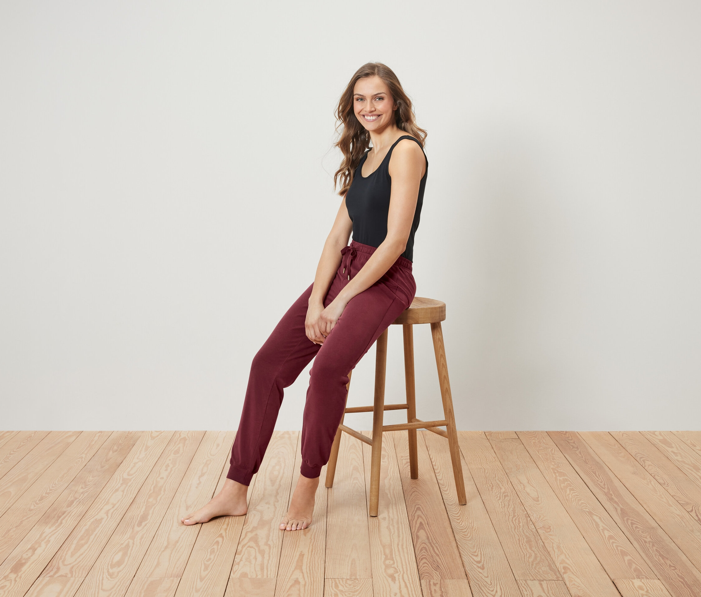 Usmívající se žena sedí na dřevěné stoličce, má na sobě černé tílko a bordó tepláky.