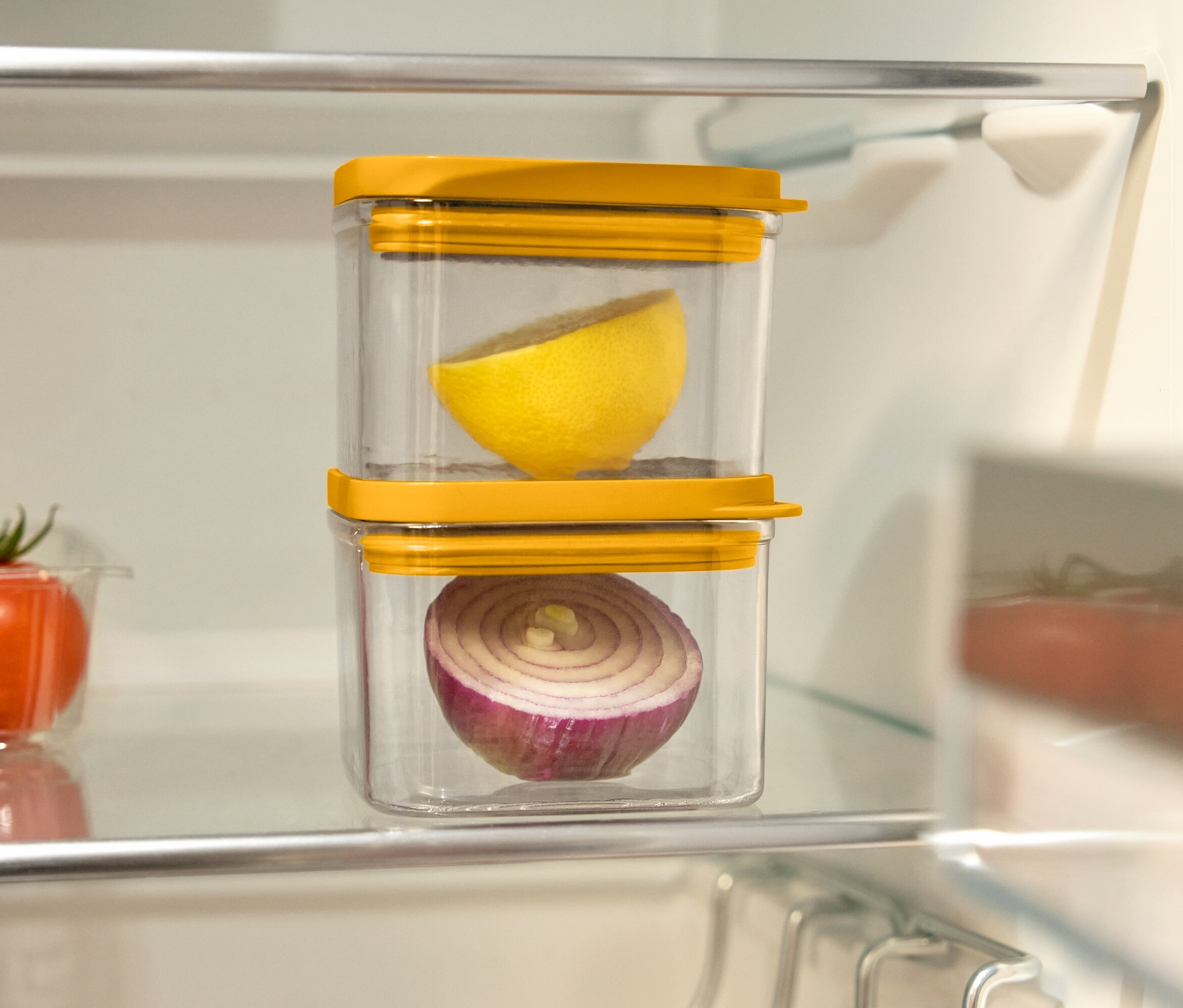 V lednici jsou dvě průhledné nádoby s žlutými víčky, jedna na druhé. V horní nádobě je půlka citronu a ve spodní půlka červené cibule. Vlevo od nich je plastová nádoba s rajčaty.