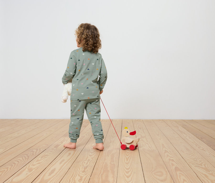 Dítě v pyžamu s potiskem srdcí tahá za sebou hračku dřevěnou kachnu na kolečkách a v ruce drží plyšového medvídka.