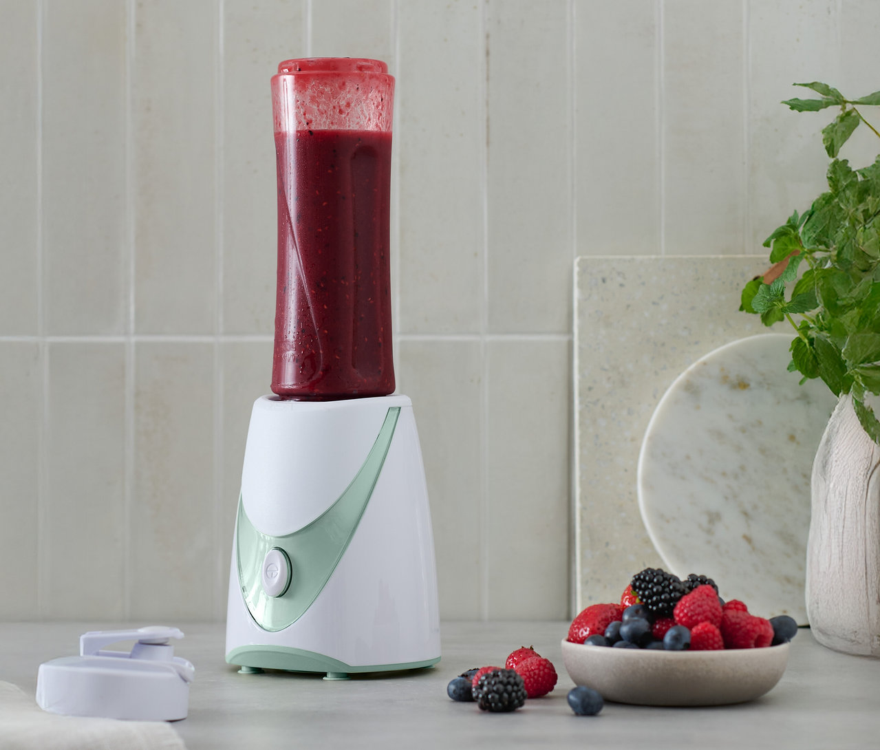 Mini-smoothie maker s hotovým smoothie na vrchu, vedle miska s bobulemi.