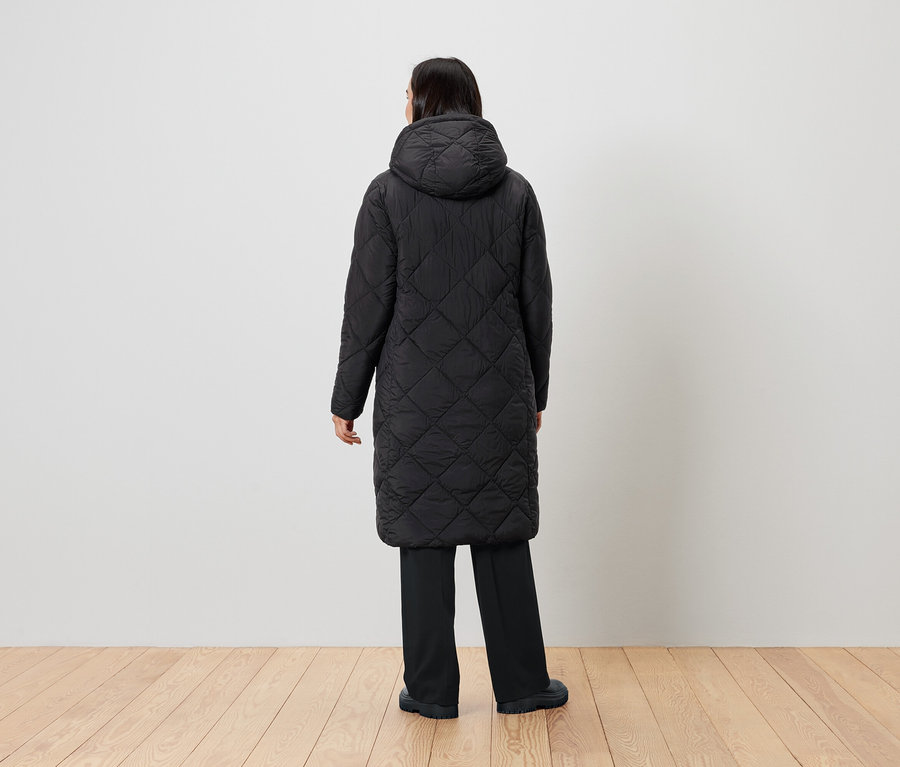 Žena stojí zády k fotoaparátu a má na sobě černý prošívaný kabát s kapucí.