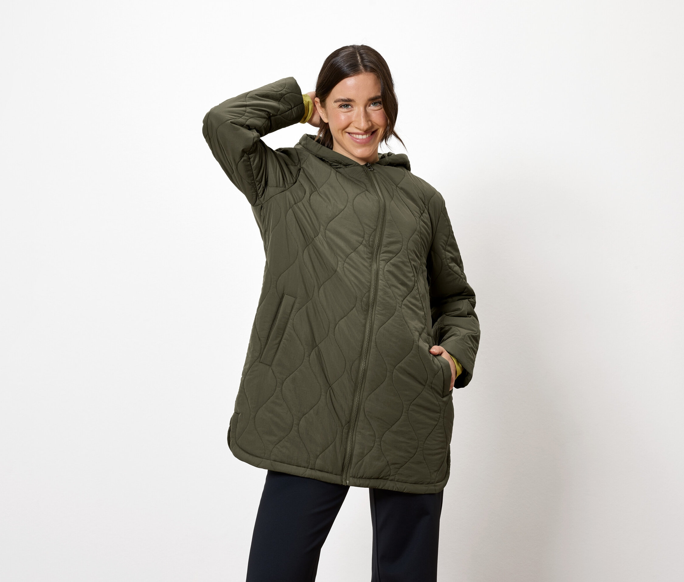 Žena pózuje v olivově zelené prošívané bundě s kapucí a tmavě modrých kalhotách.