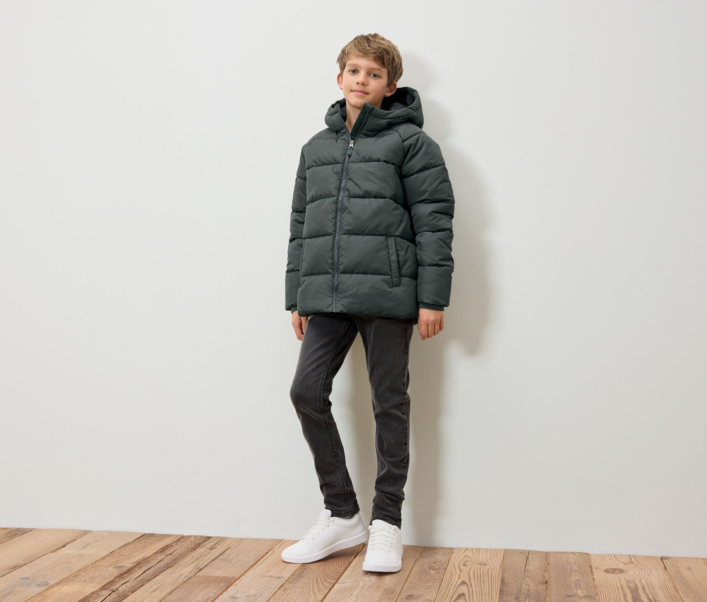 Chlapec stojí a má na sobě tmavě zelenou péřovou bundu, šedé džíny a bílé tenisky.