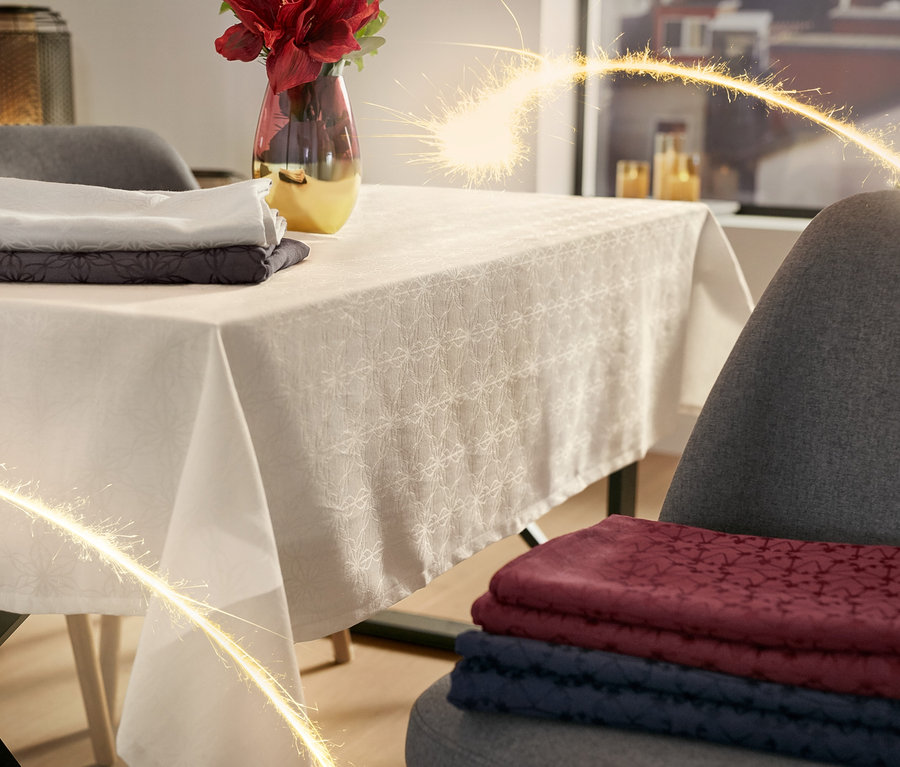 Na stole leží žakárový ubrus a váza s květinami. Na židli jsou složené žakárové ubrusy.