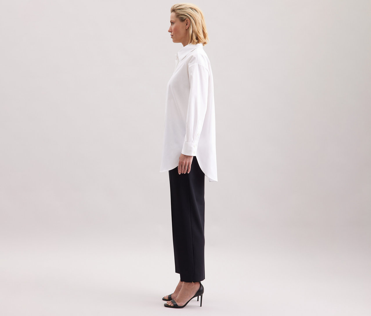 Žena má na sobě bílou dlouhou halenku oversize Seidensticker, černé kalhoty a boty na vysokém podpatku.
