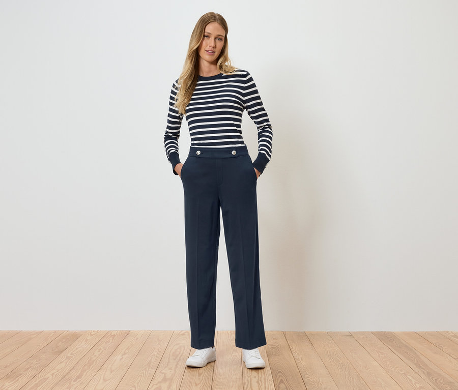 Žena stojí a má ruce v kapsách. Má na sobě pruhované tričko s dlouhým rukávem a tmavě modré kalhoty.