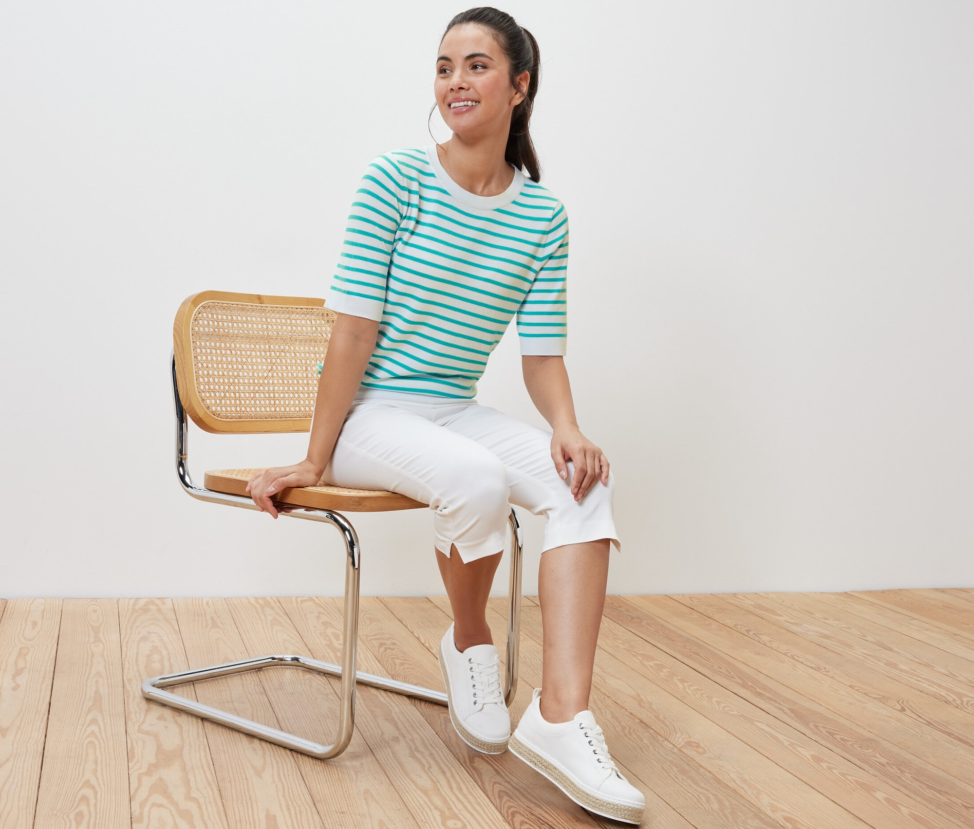 Žena sedí na židli a má na sobě modrozelený pruhovaný svetr z jemného úpletu.