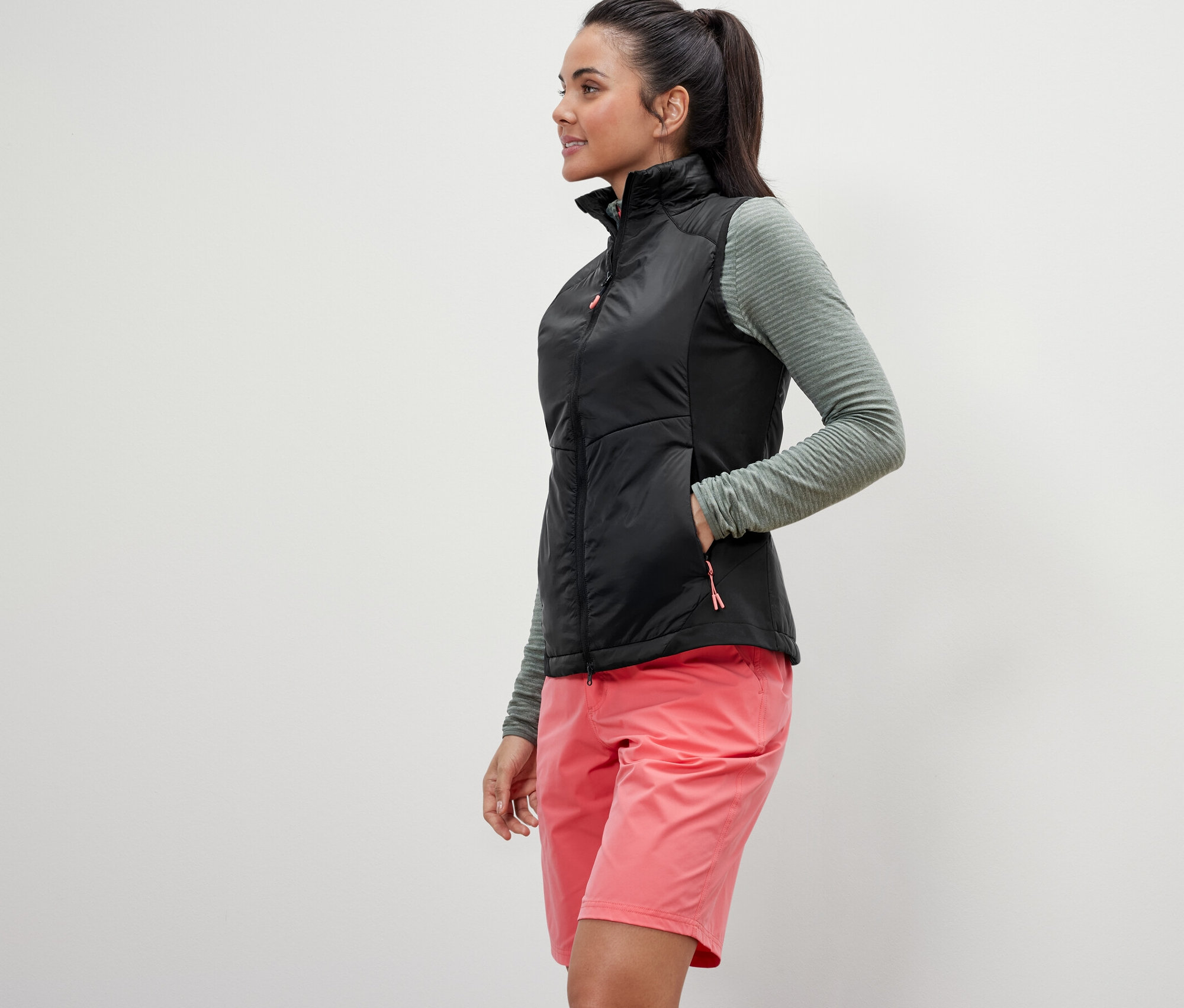 Žena pózuje v hybridní vestě »pack light«, olivovém topu a růžových šortkách.