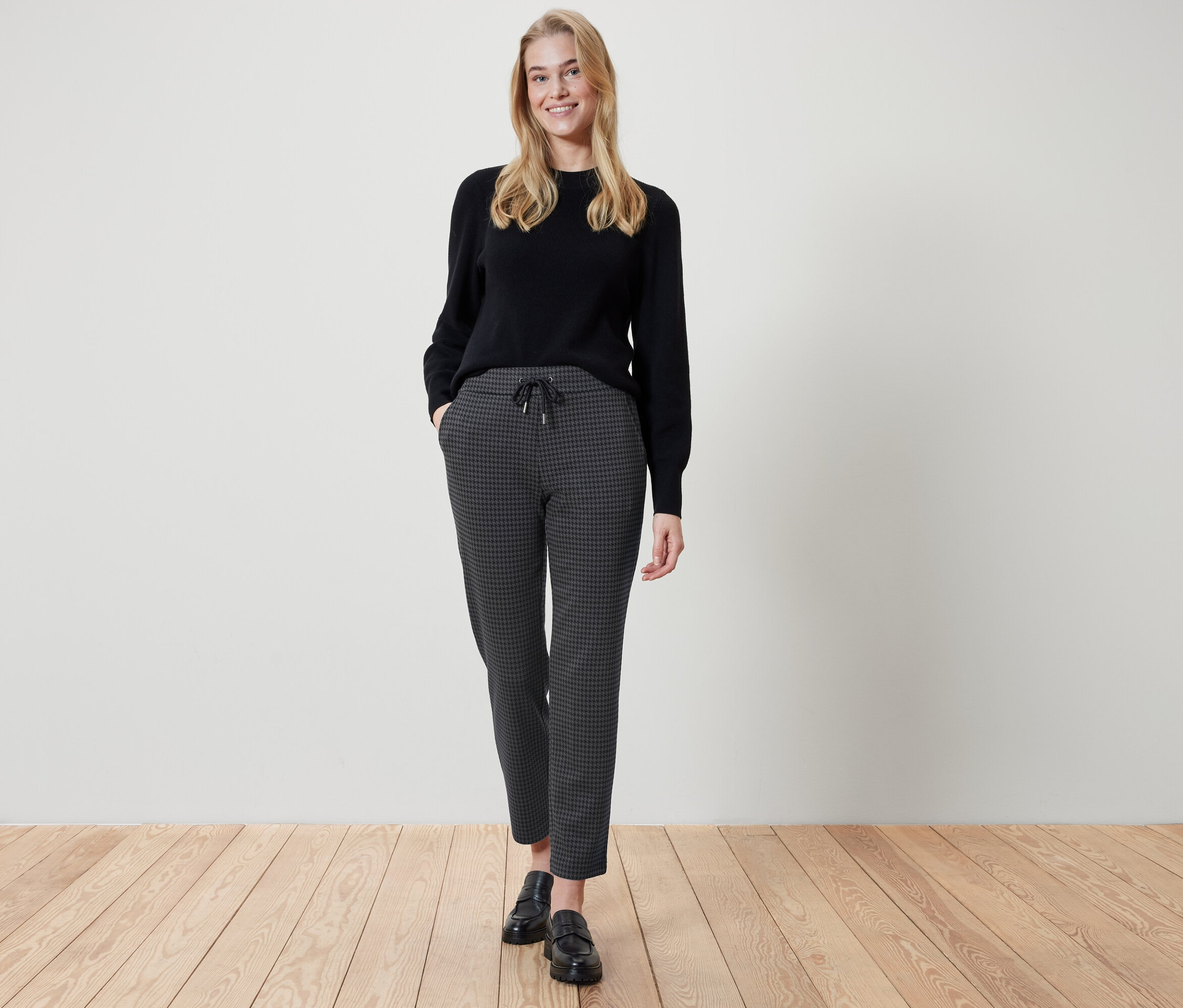 Žena stojí a má ruce v kapsách kalhot s kostkovaným vzorem. Má na sobě černý svetr a černé boty.