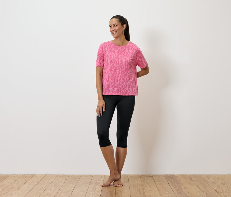 Žena stojí a má na sobě růžové tričko a černé legíny.