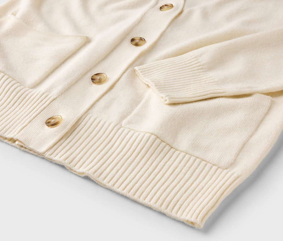 Detail krémového svetru se zapínáním na knoflíky a kapsami.