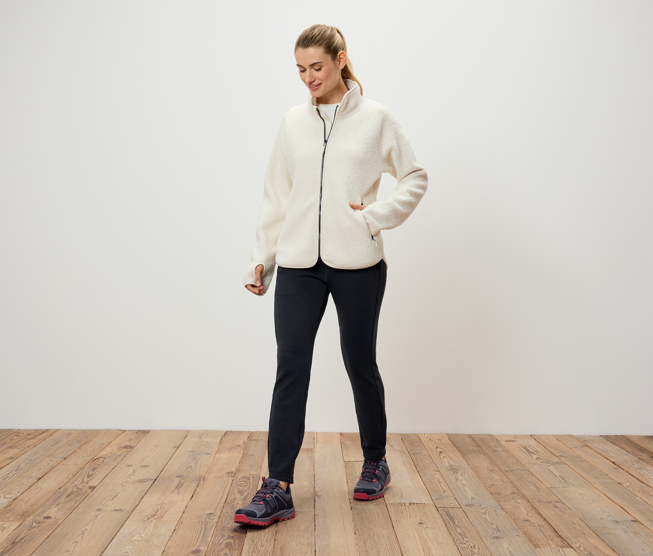 Žena v krémové fleecové bundě, černých kalhotách a sportovních botách stojí na dřevěné podlaze před bílou stěnou.