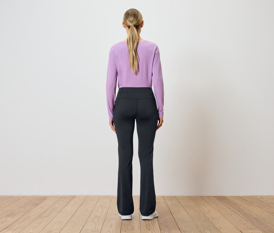 Zezadu pohled na ženu s blond vlasy staženými do culíku, oblečenou do fialového topu s dlouhým rukávem a černých kalhot.