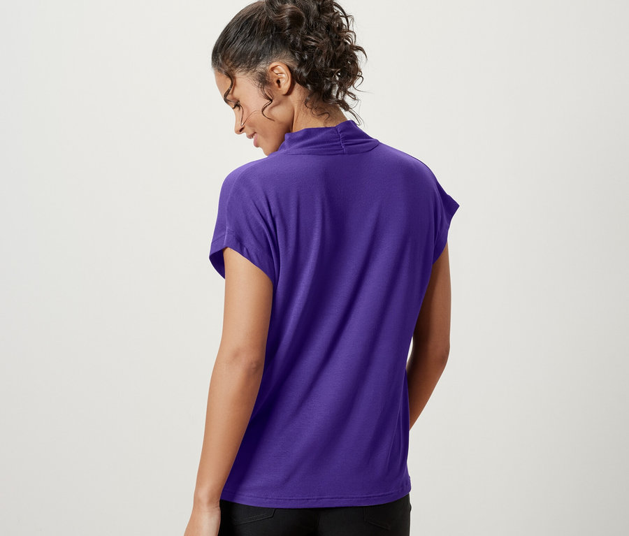 Žena zády k fotoaparátu má na sobě fialové halenkové triko s výstřihem do V a černé šortky.