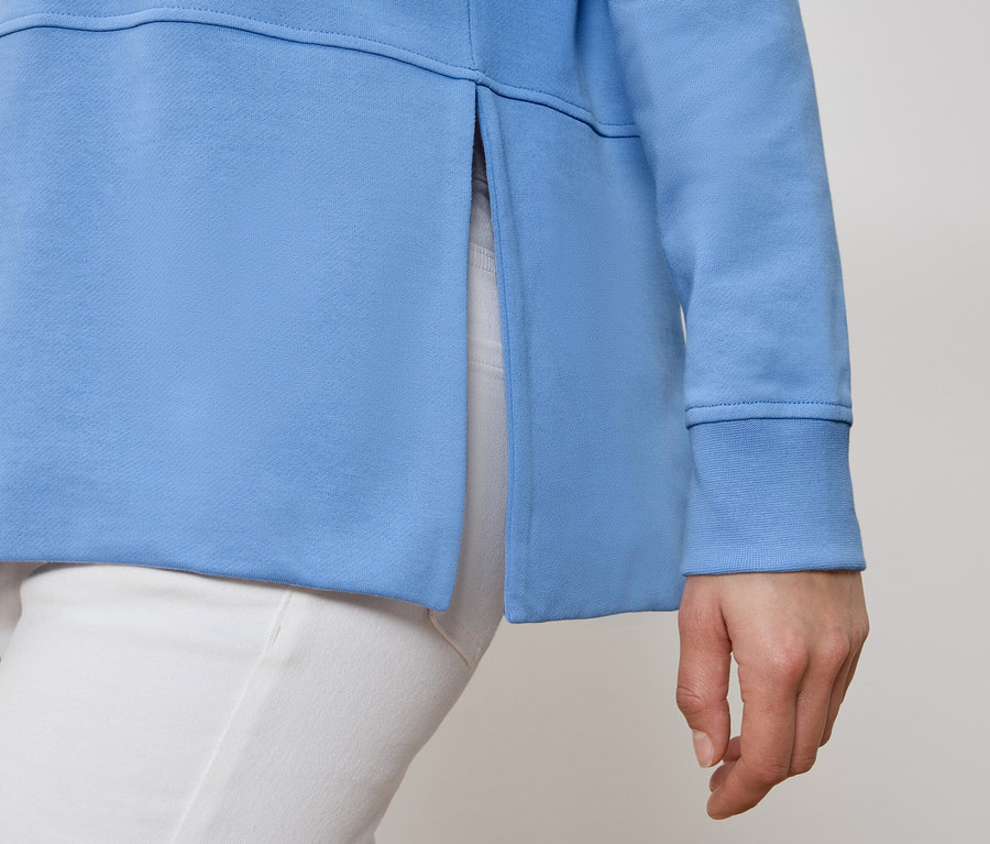 Osoba má na sobě světle modrou mikinu s bočním rozparkem a bílé kalhoty.