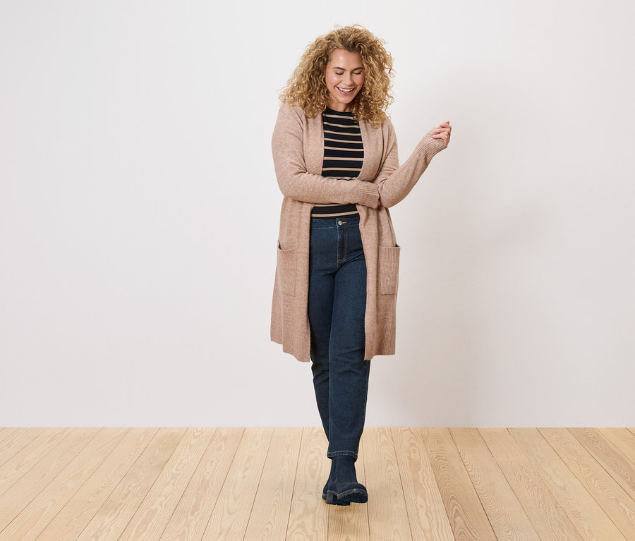 Žena s kudrnatými vlasy má na sobě dlouhý světle hnědý kardigan přes černo-bílý pruhovaný top a tmavé džíny.