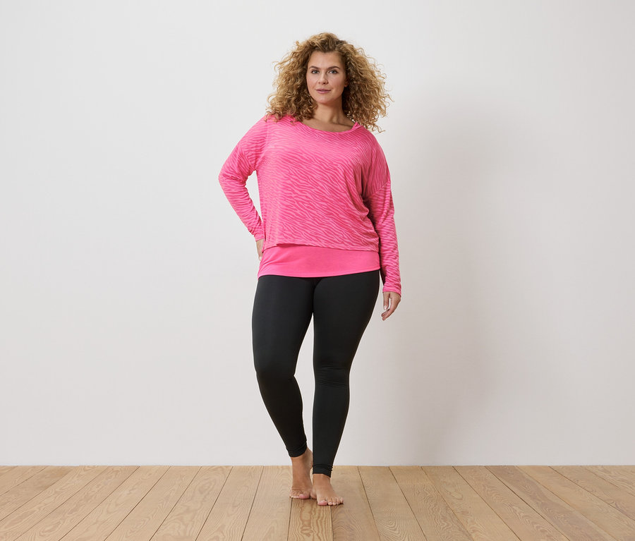 Žena s kudrnatými vlasy v růžovém zebrovaném tričku s dlouhým rukávem a černých legínách.