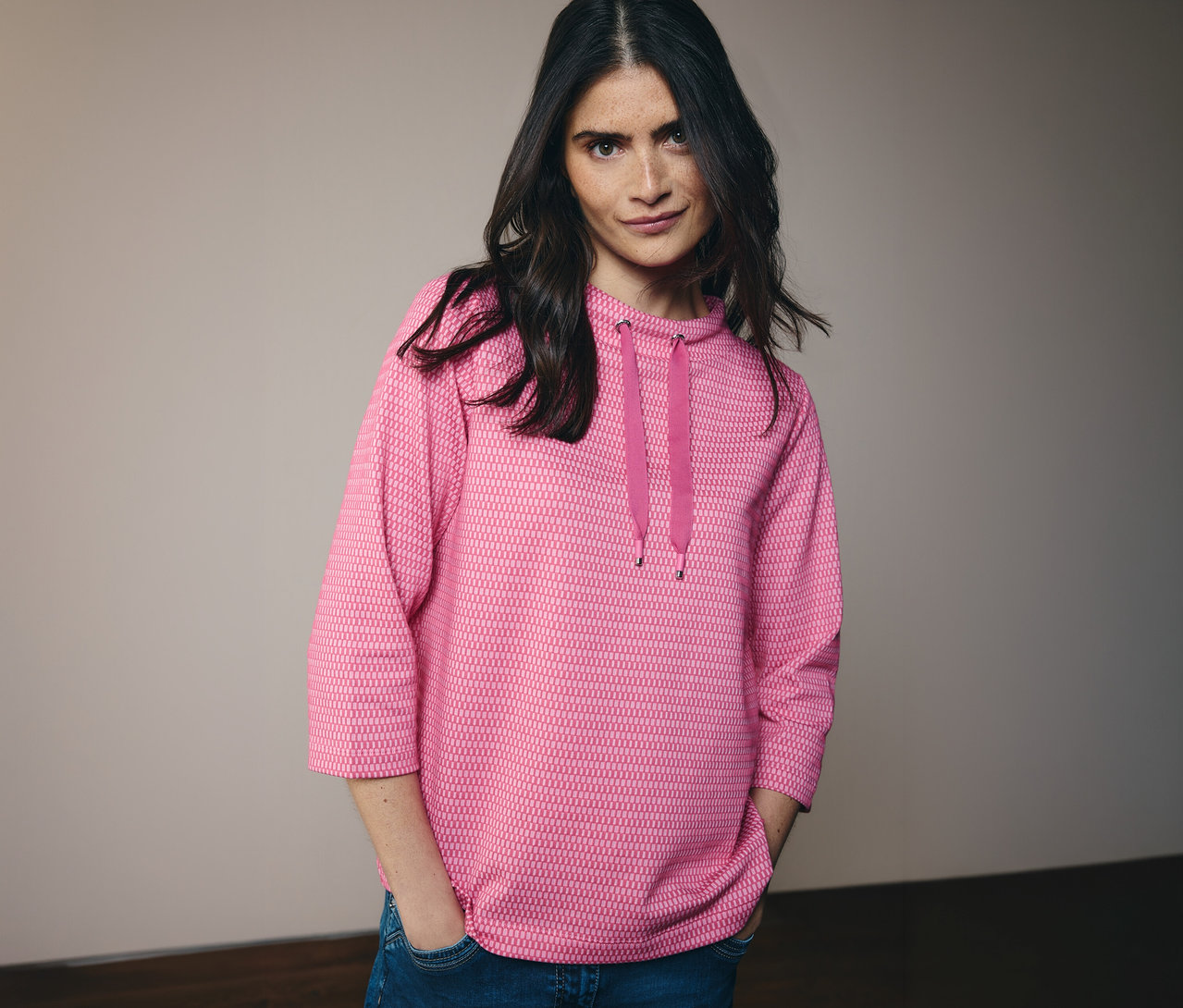 Žena v džínách a růžové mikině CECIL s kapucí a šňůrkou. Má ruce v kapsách kalhot.