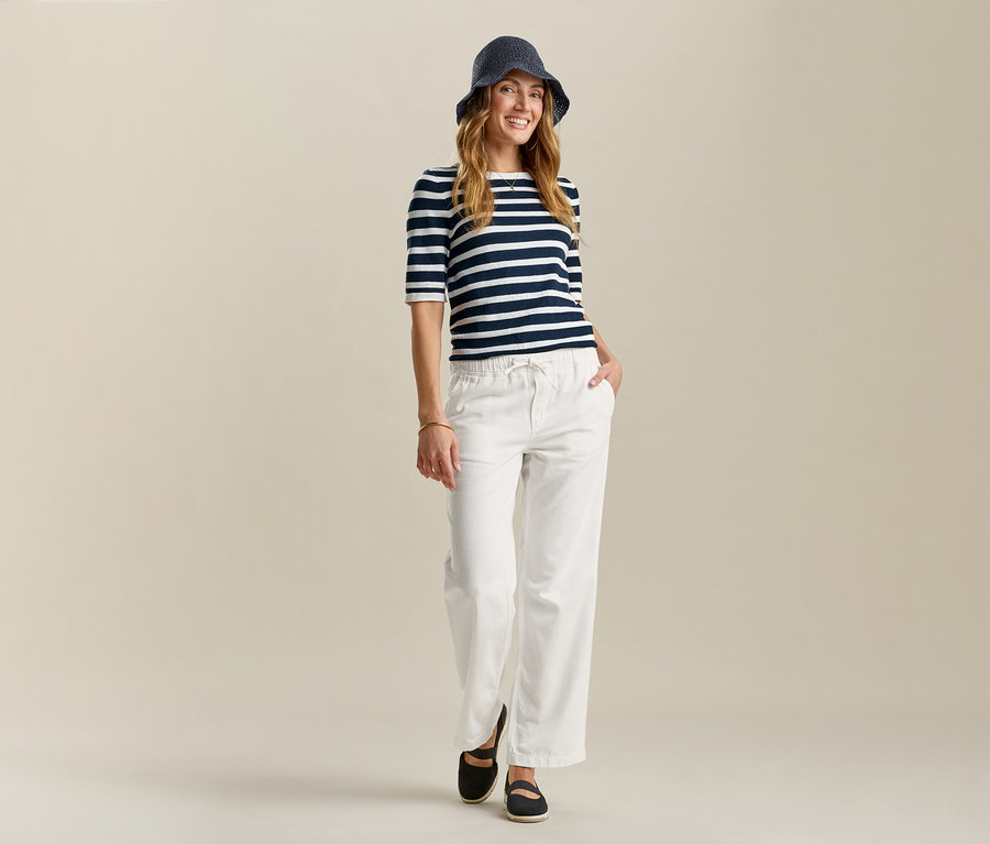 Žena v klobouku, pruhovaném tričku a Pohodlných širokých džínách pózuje s rukou v kapse na světlém pozadí.