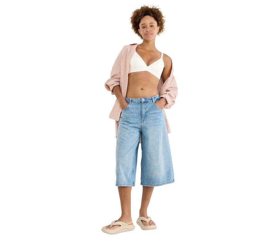 Žena stojí s rukama v kapsách, má na sobě bílou braletku, růžovou pruhovanou halenku, světle modré culottes a béžové žabky.