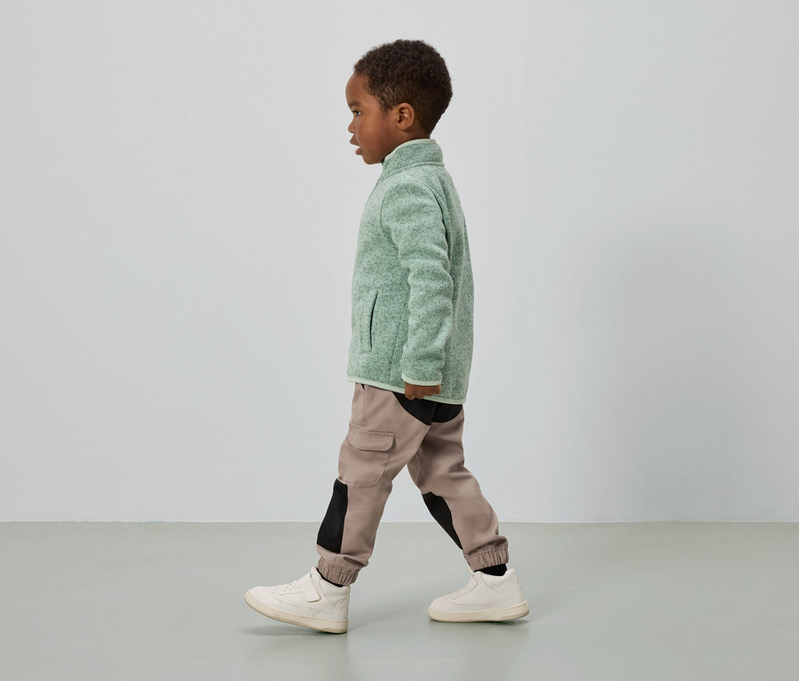 Chlapec s tmavou pletí jde, má na sobě zelenou fleecovou bundu, cargo kalhoty s černými vložkami a krémové tenisky.