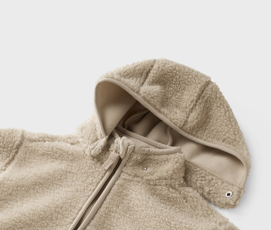 Detail béžové teddy fleecové bundy s kapucí a zipem.