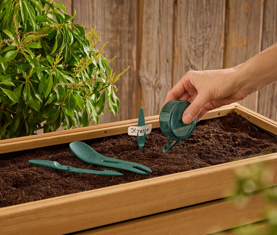 Ruka zalévá vodu z malé zelené konvičky do dřevěné bedýnky se zeminou, ve které jsou zelené zahradní nástroje a štítek s nápisem "Kresse".