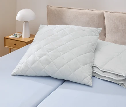 Na světle modré posteli leží bílý, prošívaný polštář Cell Solution® (90 x 70 cm). Vedle něj jsou další polštáře Cell Solution® (80 x 80 cm, 100 x 65 cm).