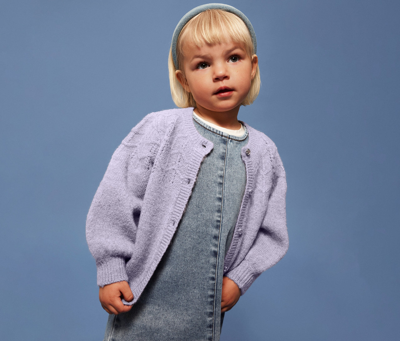 Dívka s blond vlasy má na sobě světle fialový dětský pletený svetr NAME IT přes džínové šaty na modrém pozadí.