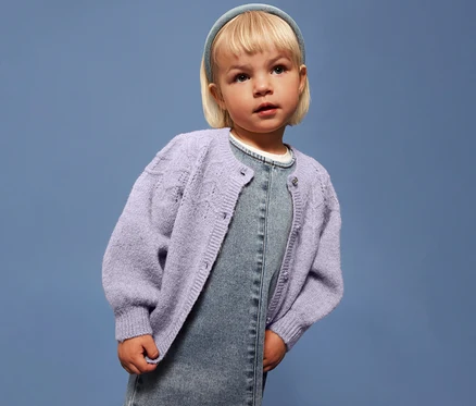 Dívka s blond vlasy má na sobě světle fialový dětský pletený svetr NAME IT přes džínové šaty na modrém pozadí.
