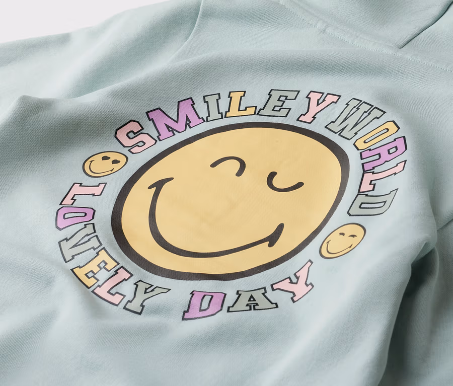 Detailní záběr mátově zelené mikiny s kapucí se žlutým potiskem smajlíka, obklopeného nápisem „Smiley World Lovely Day“.