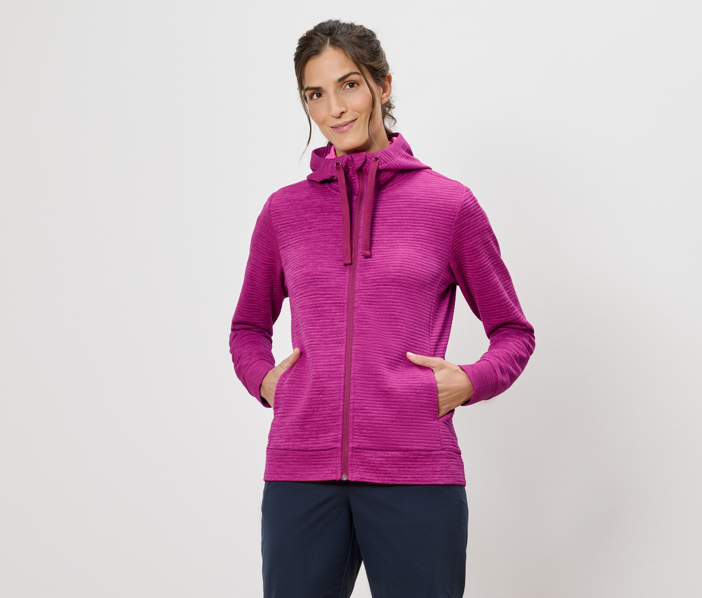 Žena v růžové mikině s kapucí s rukama v kapsách na bílém pozadí.