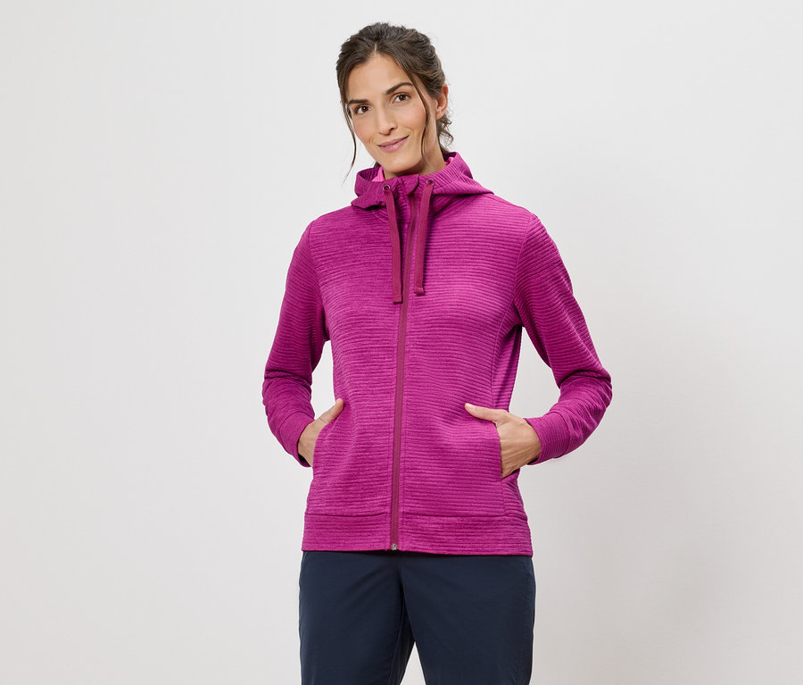 Žena v růžové mikině s kapucí s rukama v kapsách na bílém pozadí.