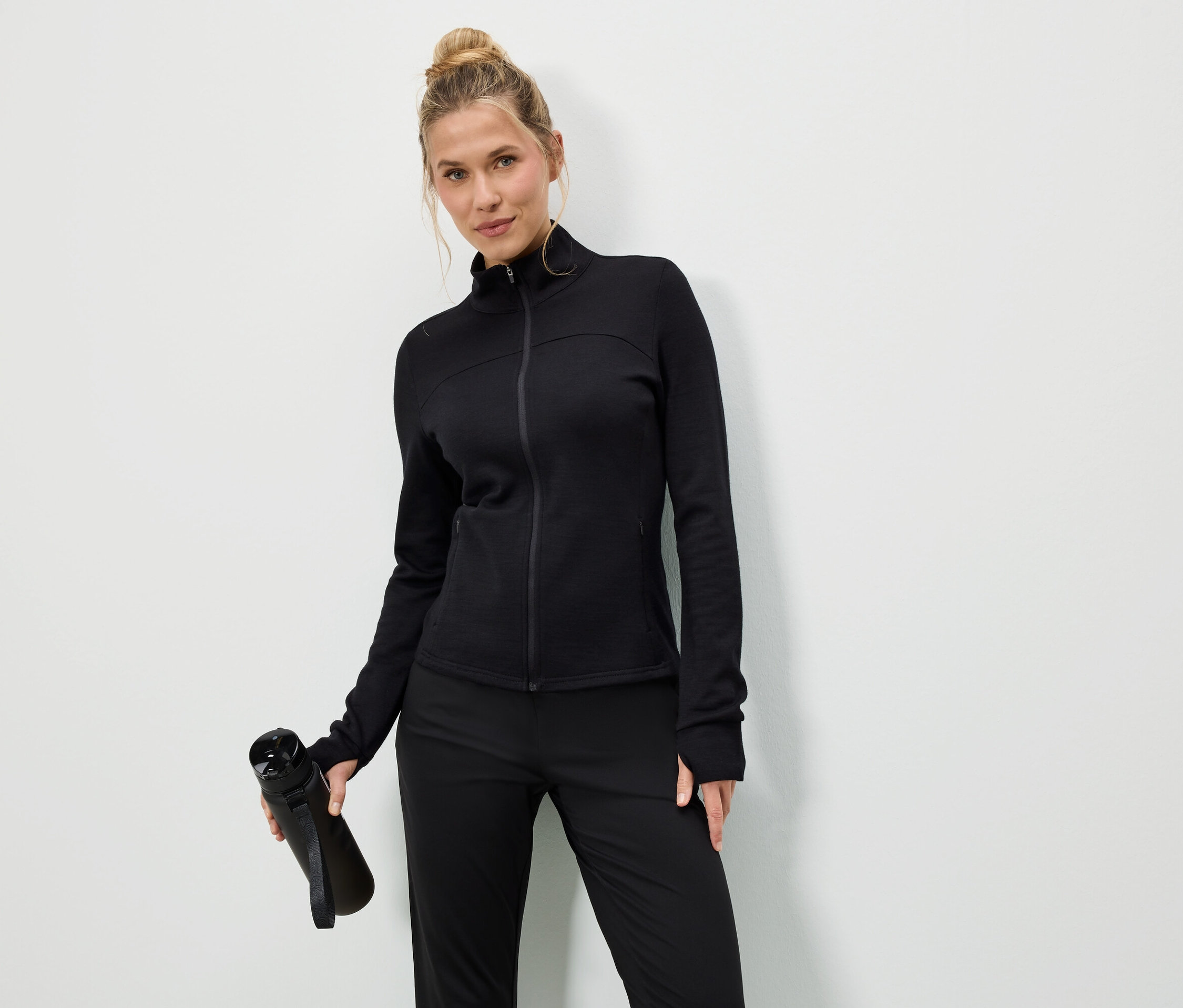 Žena v černé bundě a kalhotách drží černou láhev na vodu na bílém pozadí.