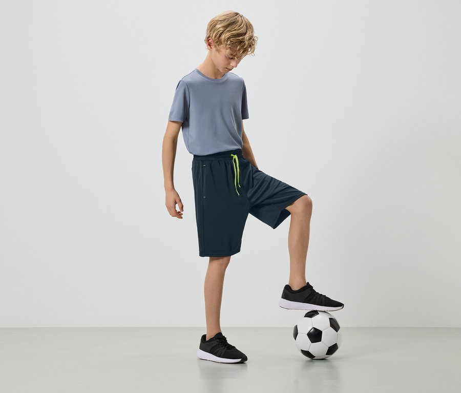 Blonďatý chlapec v šedém tričku a tmavých šortkách s nohou na fotbalovém míči.