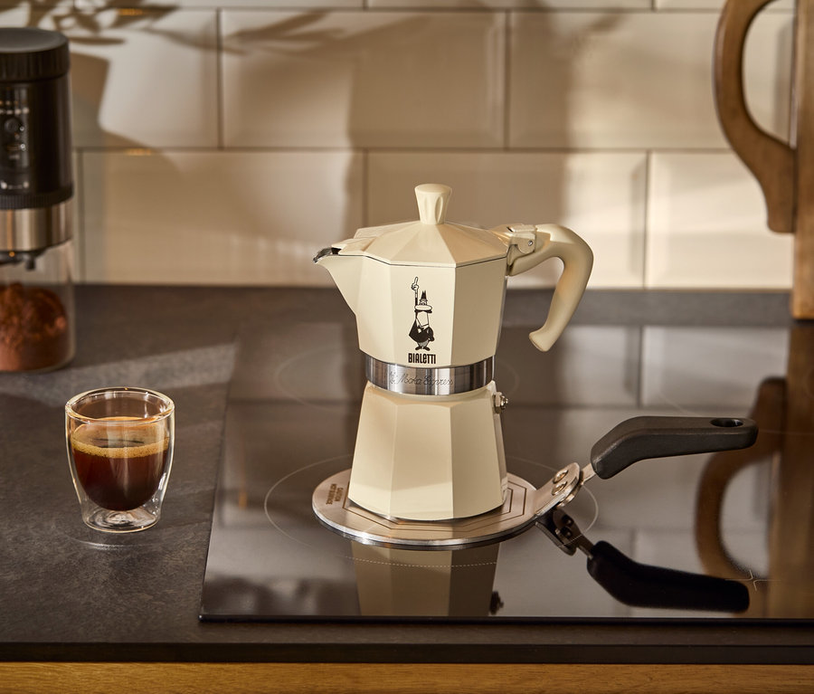 Krémová Bialetti Moka Express, 190 ml stojí na indukční plotýnce Bialetti na varné desce. V pozadí je šálek kávy.