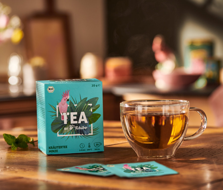 Balení BIO TEA by Tchibo máta vedle šálku čaje a lístků máty na dřevěném stole.